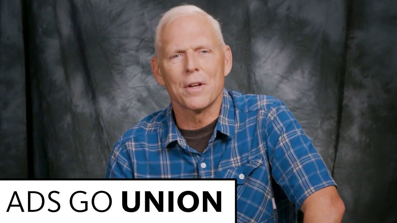 Actor in blue button up with grey shirt underneath speaking into camera with black and grey tie die background