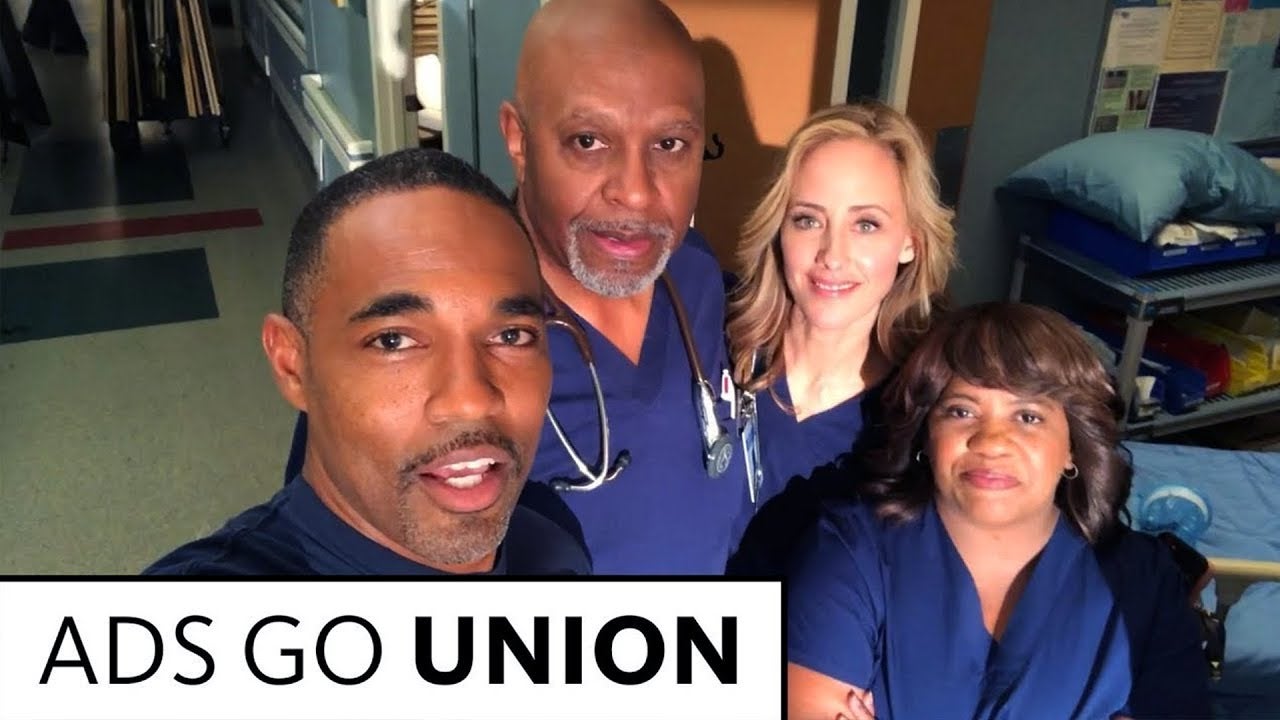 Grey's cast in dark blue scrubs looking up to camera being held by George on left. 