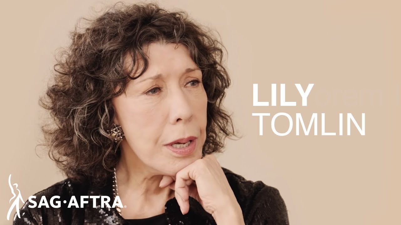 Tomlin in black top gently rests chin on left hand knuckles. Tan background with 'Lily Tomlin' in white off to right and SAG-AFTRA logo in bottom left corner