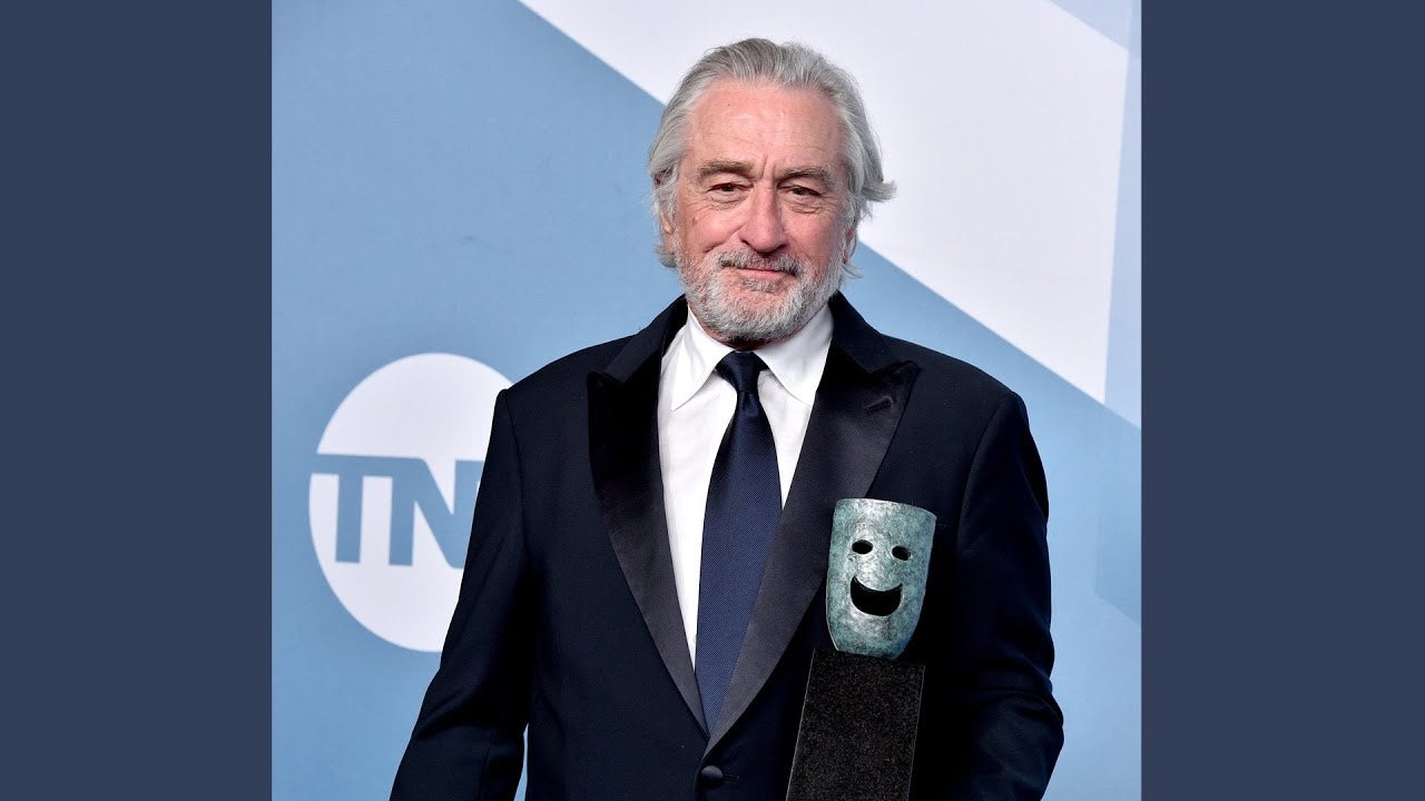De Niro n white button up, navy blue tie and black jacket standing behind SAG Life Achievement Award in front of blue SAG Awards, TBS and TNT step and repeat