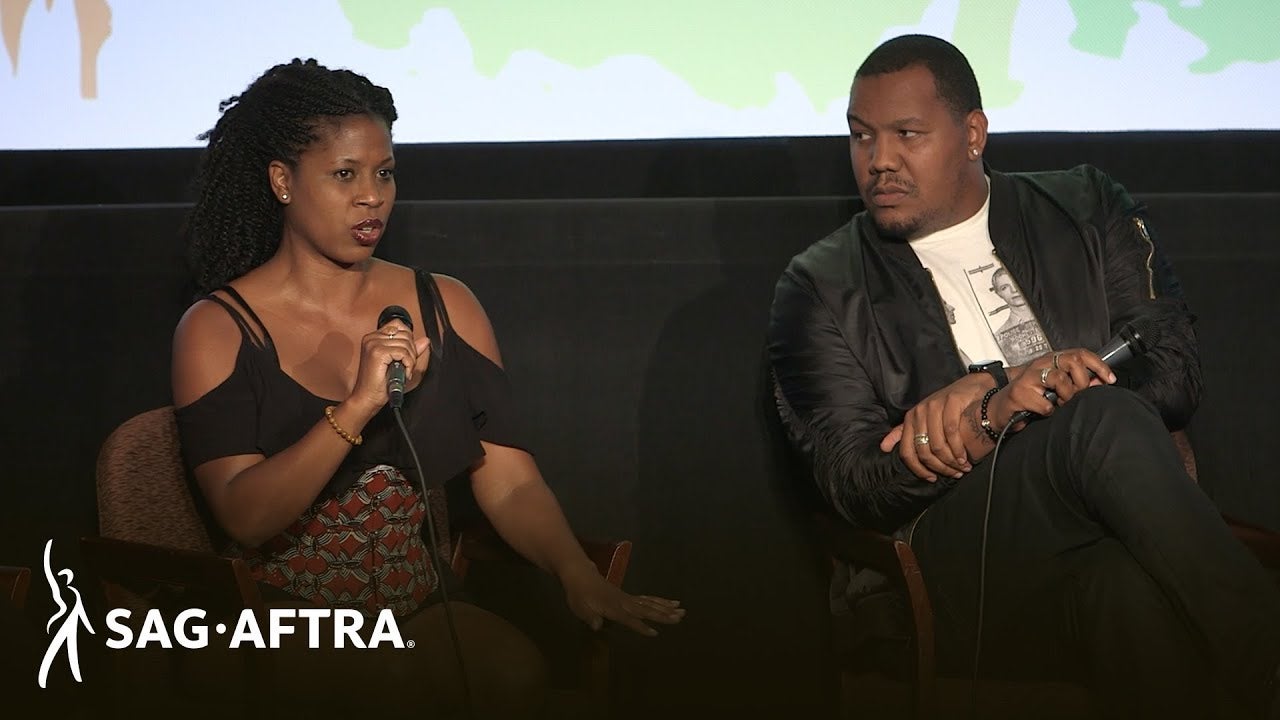 Female panelist on left in black top holding mic in left hand speaking and looking out at audience. Male panelis to her left wearing a white shirt and black jacket sits cross-legged holding mic in left hand looking at panelist to his right.