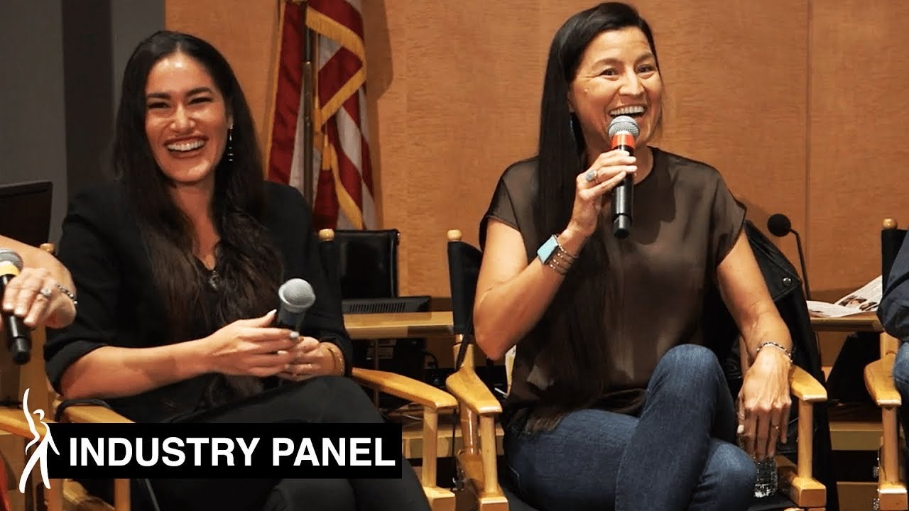 Two female panelists sitting in Directors Chairs holding microphones.Panelist on left smiling at the audience while panelist on right speaks into the mic looking out at the audience