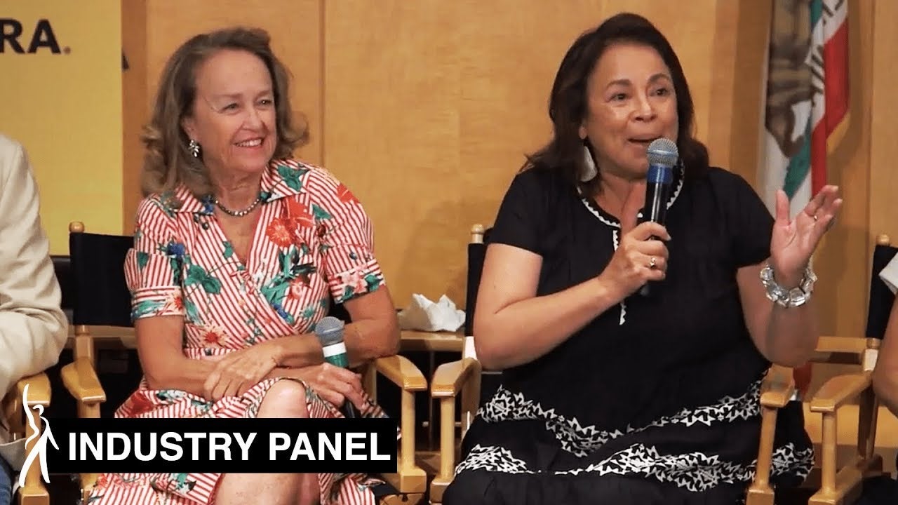 Two female panelists sitting side by side in director chairs. Panelis on left looking over at panelist on right speaking into her mic, looking at the audience.