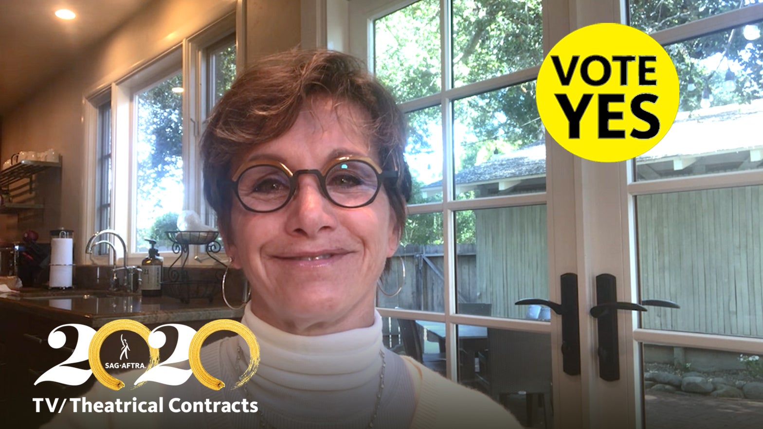 Carteris in white turtle neck and circle glasses in front of windows with a yellow "Vote YES" with black text in the top right corner and 2020 TV/Theatrical logo in bottom right corner
