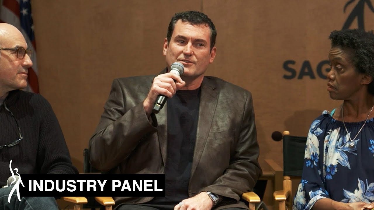3 panelists in still image. Male panelist on left looking over at panelist in the middle. Male panelist in middle speaking into microphone in right hand looking out at audience. Panelist on right looking at middle panelist speaking.