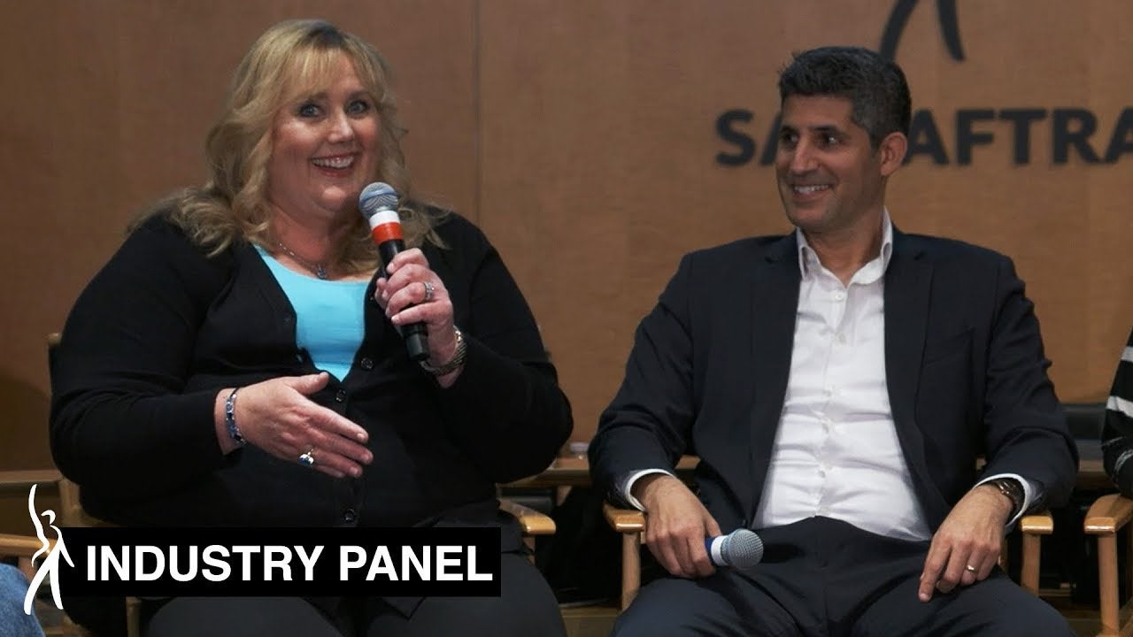 Female panelist on left speaking and smiling into mic looking out at audience. Male panelist on left holding mic in right hand smiles at panelist on left.