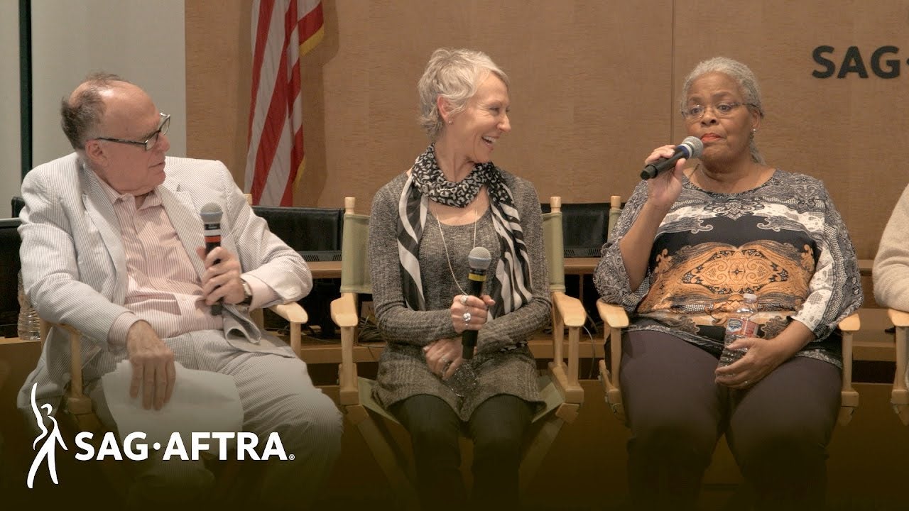 "Panelist on left leaning to right looking at panelist on far right. Panelist in middle wearing grey longsleeve top with black and white scarf looks over at panelist on right. Panelist on right in colorful patterned top speaks into mic in right hand