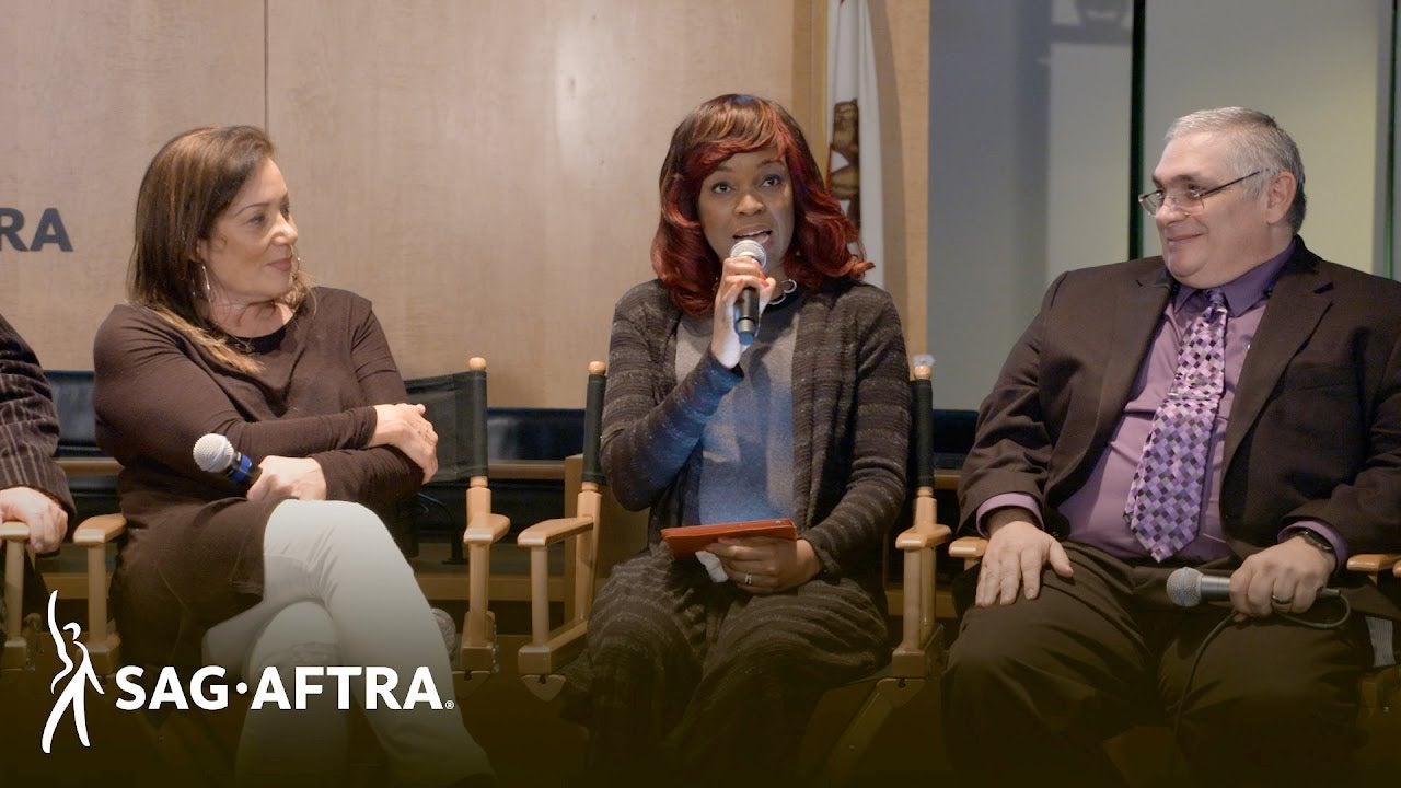 Panelist on left in brown with arms crossed holding mic, middle panelist looking out at audience speaking into mic in right hand and panelist on right in red and blue plaid button up and brown blazer looing to his right at panelist speaking
