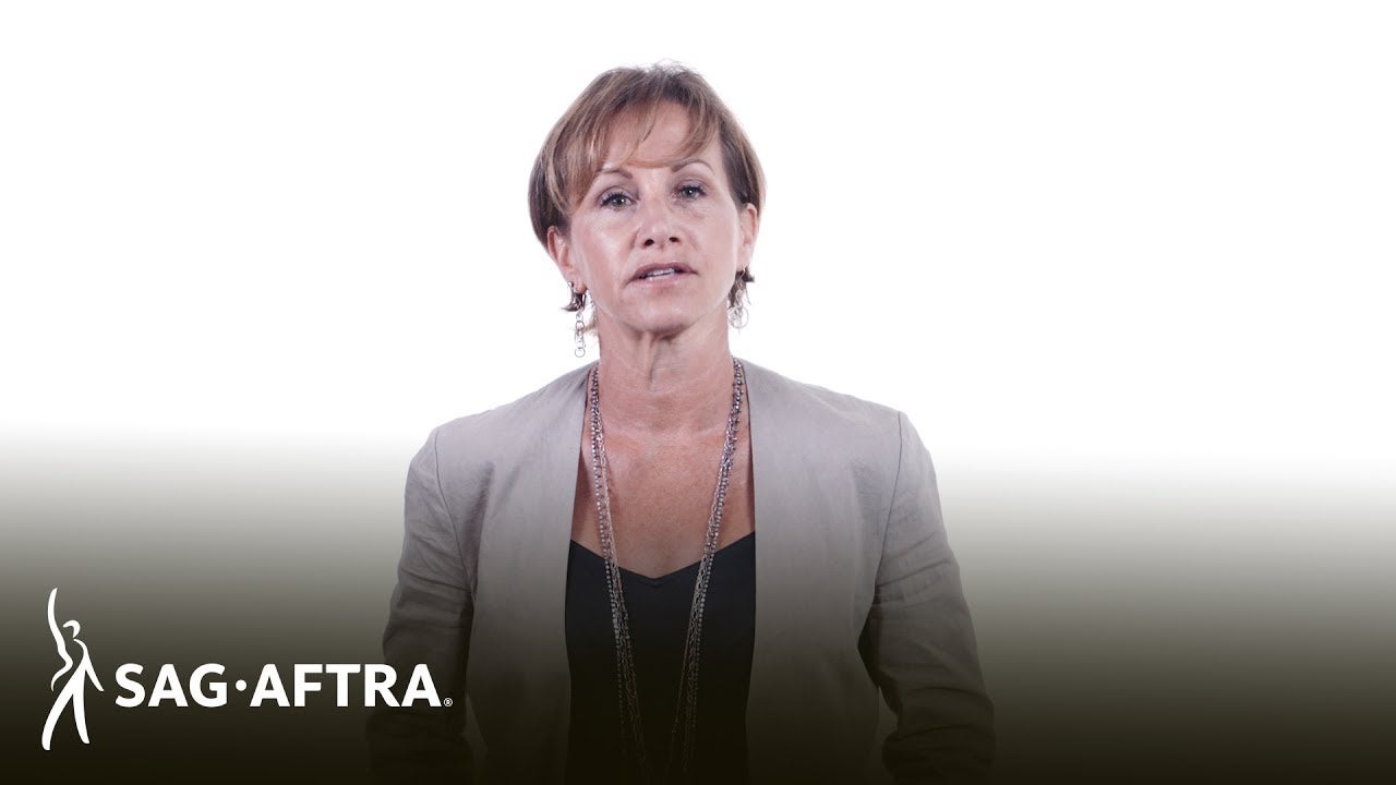 Carteris in dark top and beige blazer and long necklaces standing in front of a white background