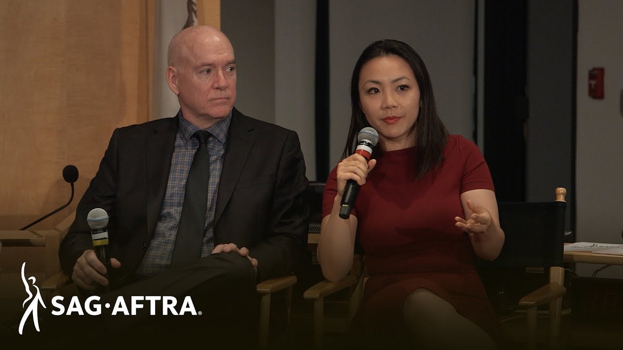 panelist on left in grey button up, black tie and black blazer holding microphone in right hand looking at panelist on right. Panelist on right in red dress speaking into microphone in her right hand looking out at audience