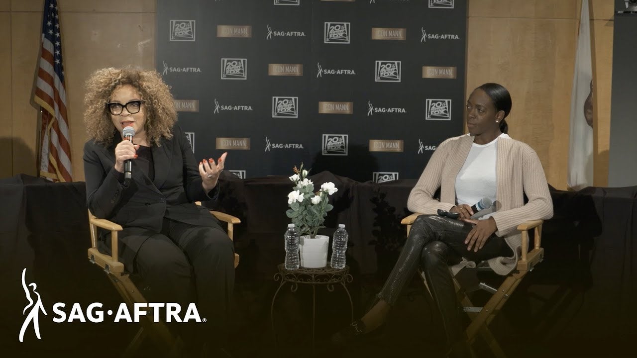 Panelist on left in all black suite looking out at audience with panelist on right in white top and beige cardigan and black leggings