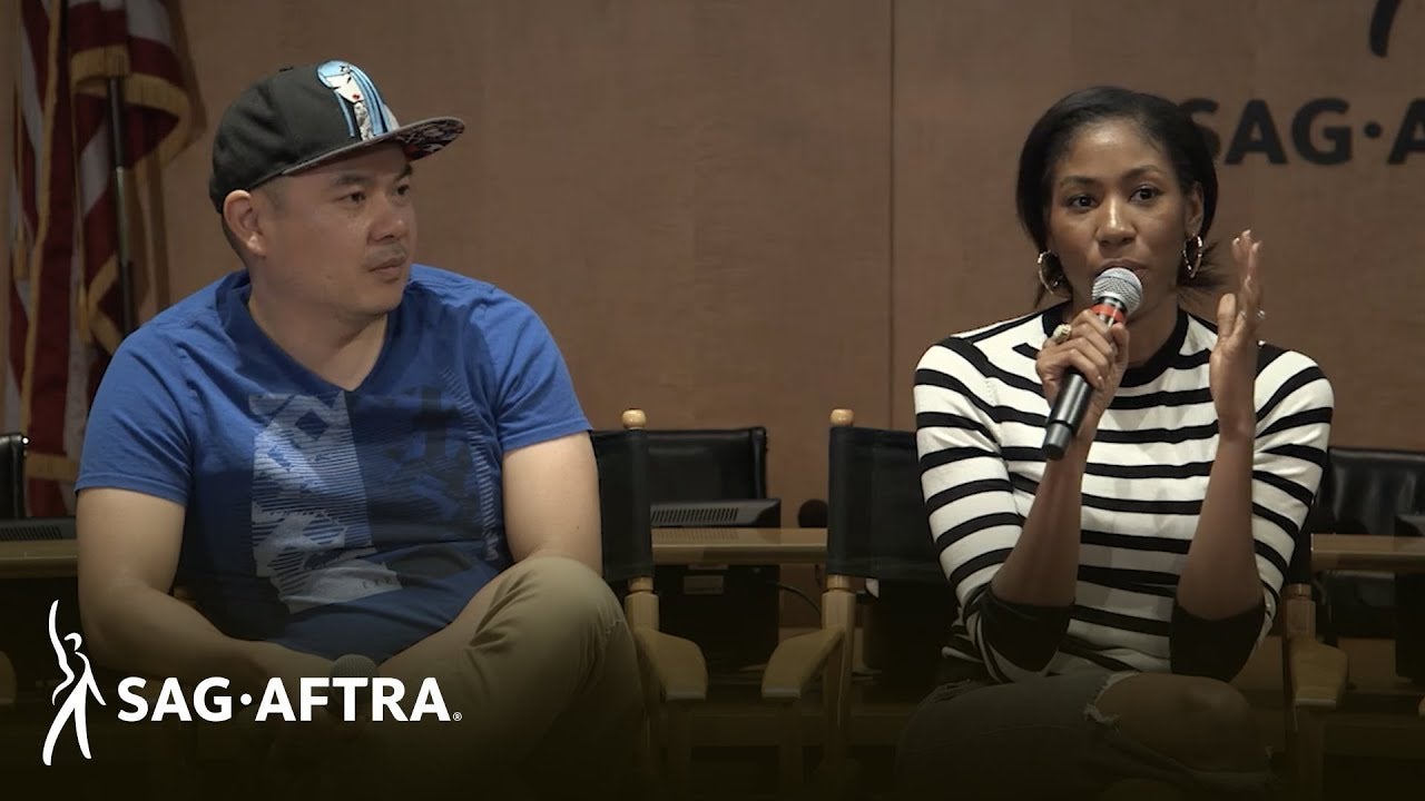 Panelist on left in blue shirt and black hat on left looking out at audience and panelist on right speaking into mic in right hand gesturing with her left wearing a black and white horizontal stripped top