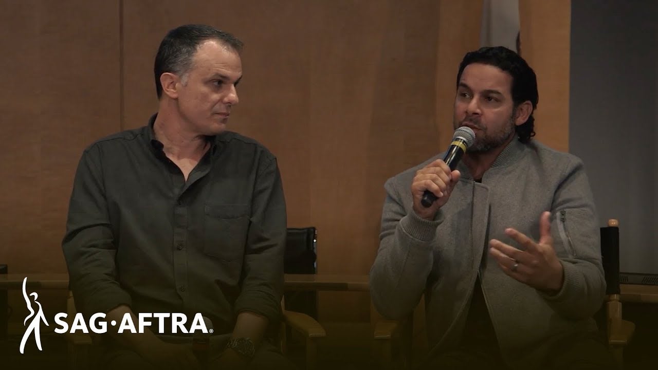 panelist on left in black button up looking over at panelist on right speaking into microphone looking out at audience