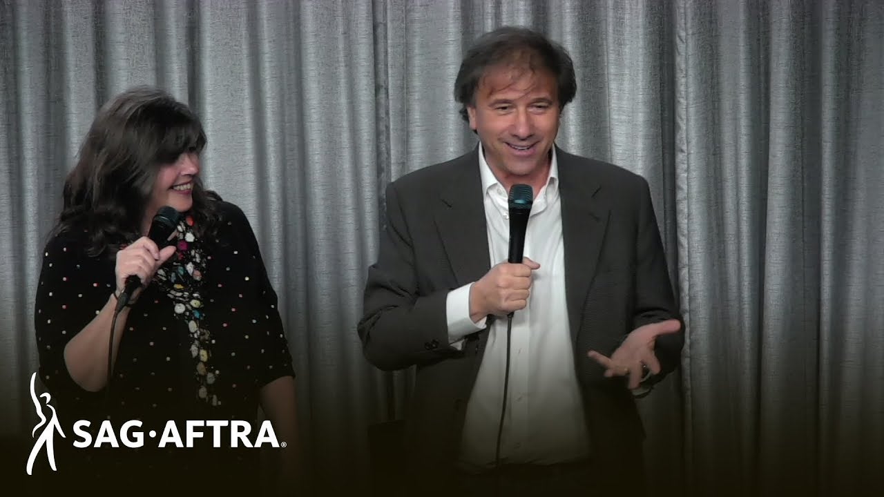 Megan on left holding mic in right hand looking at Chuck on right in white button up and grey blazer speaking to audience