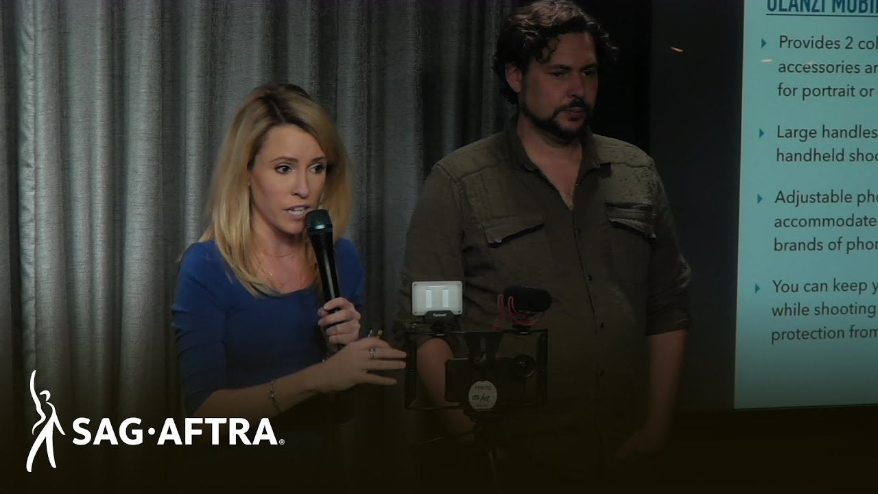 Mozino in blue U-Neck top holding mic in left hand with tripod and mobile device in front of her. Mestman to Mozino's left