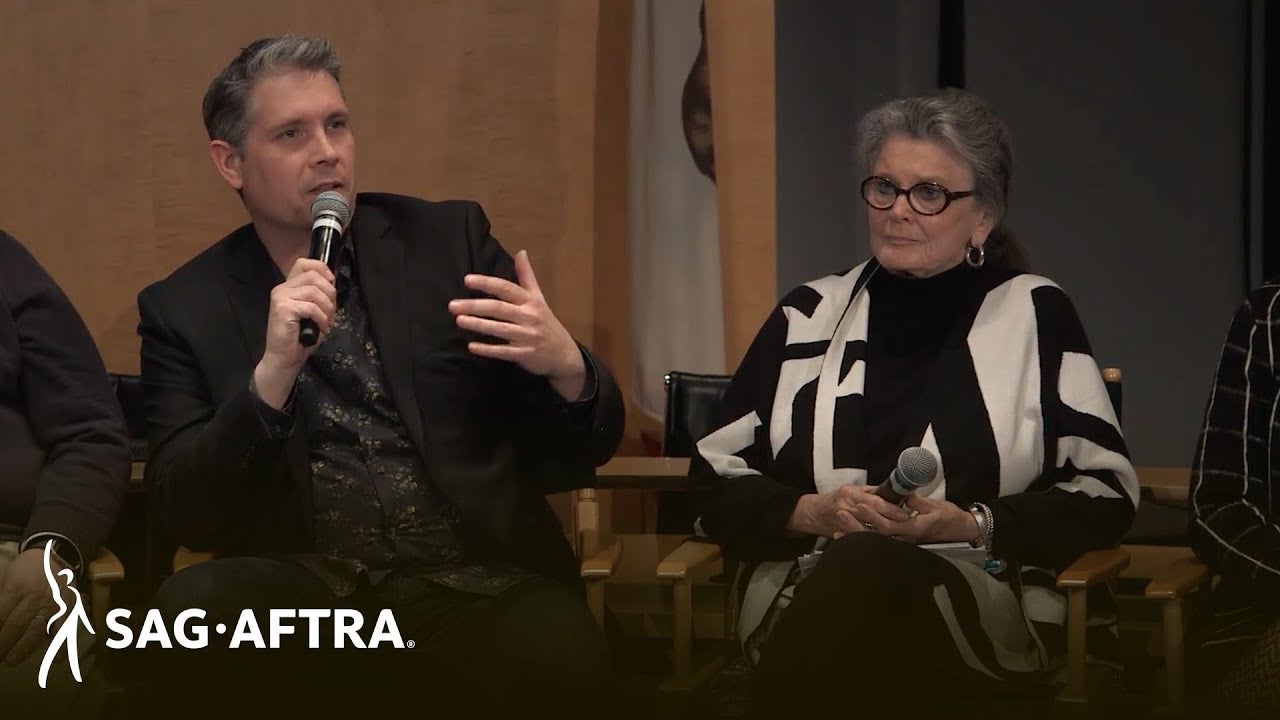 Panelist on left in dark shirt and black blazer speaking into microphone looking out at audience. Panelist on right in black top and black and white cardigan looking to her right at panelist speaking.