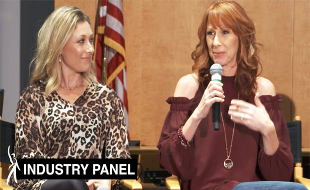 Two panelists sitting side by side. Panelist on left looking to her left at panelist on right speaking into a microphone.