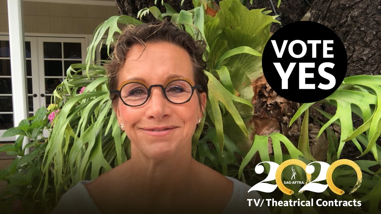 Carteris in front of green foliage smiling. "VOTE YES" in white text in a black circle in the top right corner and White 2's and gold brush stroke 0's written out in 2020 with "TV?Theatrical Contracts" in white below in bottom right corner