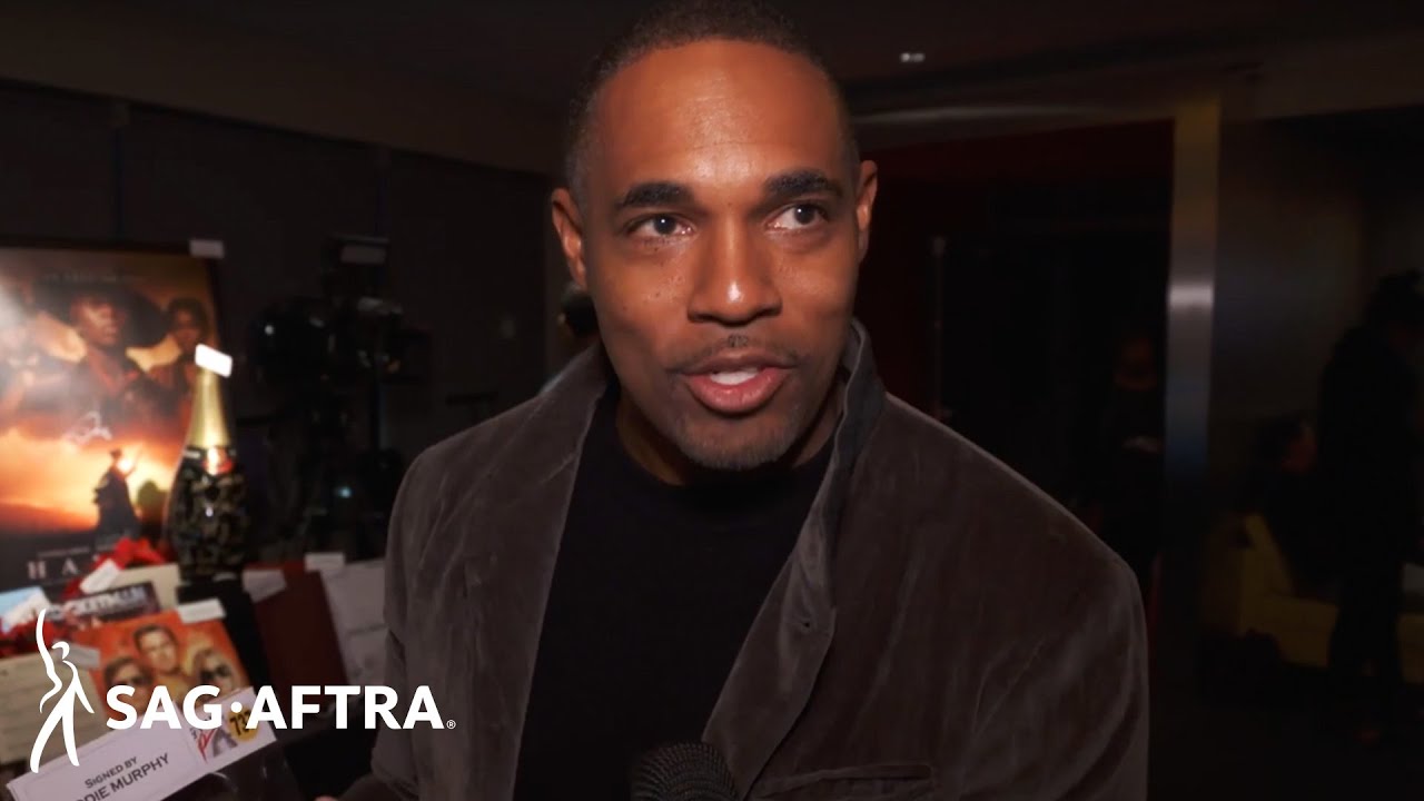 Jason George in black shirt with dark brown velvet blazer looking slightly off camera with auction table in background.