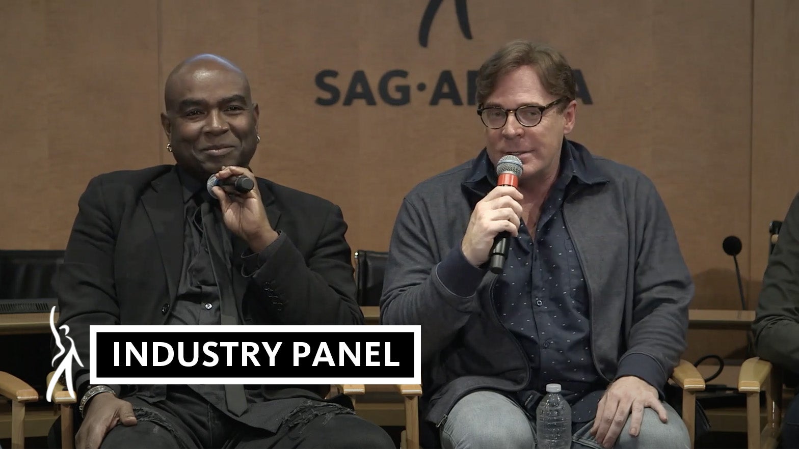 Panelist on left in all black suite holds mic towards chin looking at audience while panelist on the right speaks into the microphone in his right hand looking out at the audience.