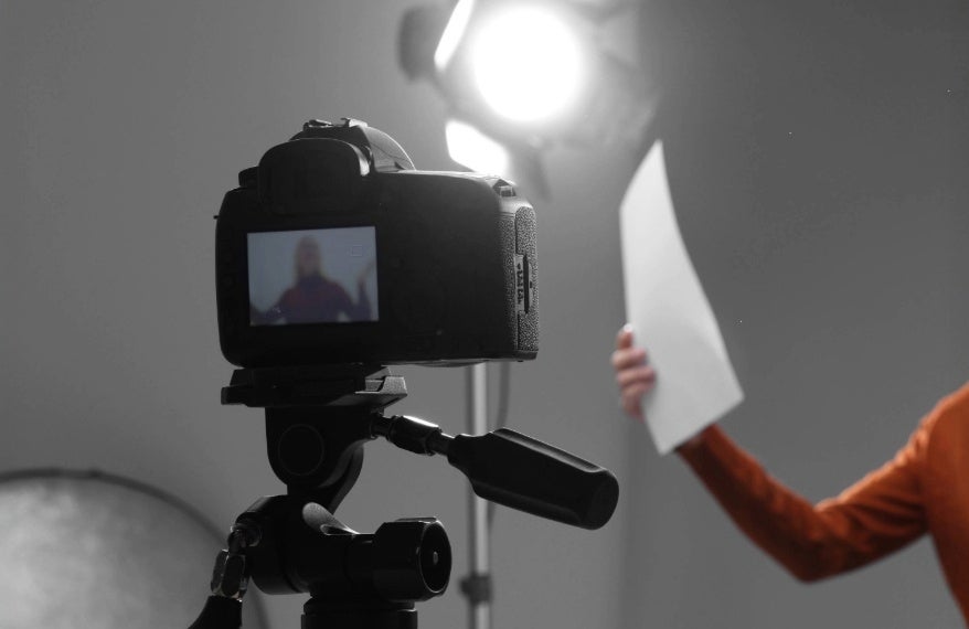 An image of a black camera on its tripod filming a woman holding a piece of paper in her hand with a light shining on her.
