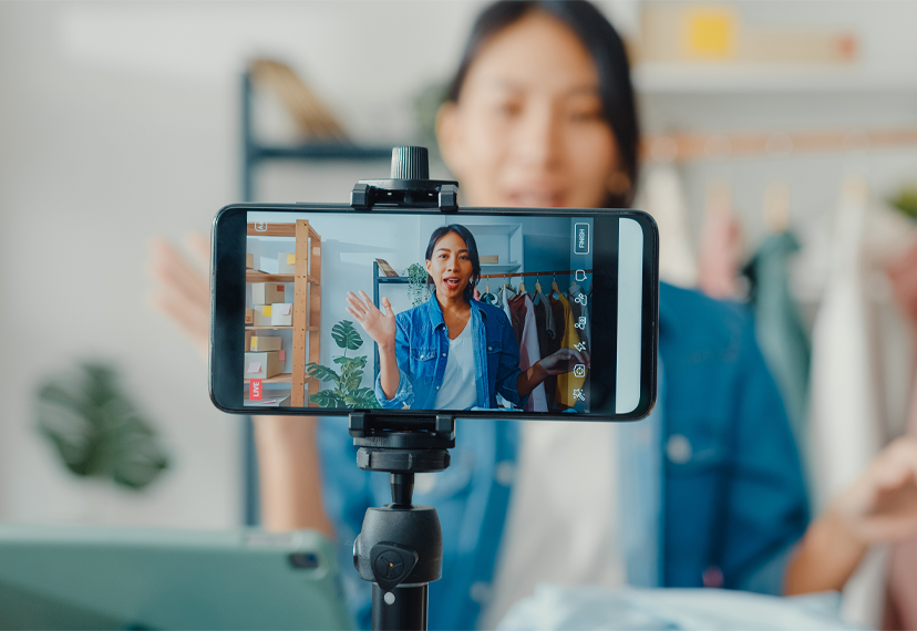 A young woman recording a video on her smartphone, which is mounted on a tripod. She appears to be explaining something, with a clothing rack and home decor in the background.