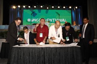 DCI in suite next to Gabrielle Carteris leaning over a table, Ochoa pointing to the crowd with his righ thand, Treviño in white leaning over a table signing papers, Calva leaning over table in a black shirt and DW in a suite
