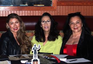 Three women sit at a table. On the table are SAG Awards scorecards and a cutout of The Actor statuette.