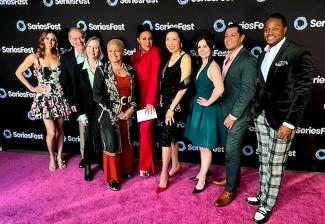 A well-dressed group of nine people stand on a fuchsia carpet in front of a backdrop with multiple SeriesFest logos.
