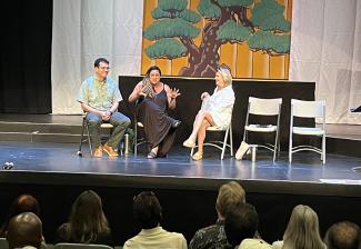 Three people, one man in an aloha shirt, one woman in a black sundress, and a woman in a white shirt dress, sit on a well-lit stage before an audience of actors. There is a backdrop with an illustration of a bonsai tree.