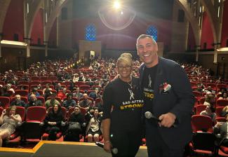 Two individuals stand on a stage in front of an audience of people in a theater.