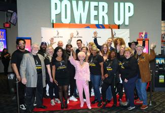 A group of men and women pose with their hands raised high cheering in front of a SAG-AFTRA backdrop. 