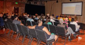A view from behind a few dozen people, who sit in chairs listening to four women, who are seated at a yellow table at the front of the room. Behind them is a screen. 