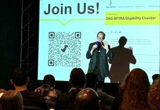 Gleason, dressed in a dark blazer over a SAG-AFTRA T-shirt, stands in front of a projector screen with the words “Join Us - SAG-AFTRA Eligibility Tracker” and a QR code. Seated attendees can be seen in the foreground.