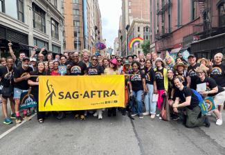 A large group of people gather behind a yellow parade banner with a black SAG-AFTRA logo in the center. The people wear black T-shirts with a rainbow and white SAG-AFTRA logo in the center. Some hold teal fans with a rainbow and white SAG-AFTRA logo.