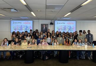 A diverse group of people smile in front of a long tan table. Two screens that read Kids and Teens Day at the Sag-Aftra Foundation are on either side of them.