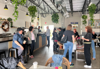 People mill about in a coffee shop, one on a stool, others sitting at tables and one leaning on the counter. Plants hang from the ceiling. 