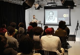 Looking toward the front of the classroom from the back. An instructor stands next to a screen, while attendees are seated.