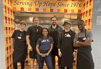 Six people stand in an alcove adorned with colorful tiles. Above the doorway, it reads "Serving Up Hope Since 1976." All the people wear aprons except for the woman in the center, who wears a dark blue T-shirt.