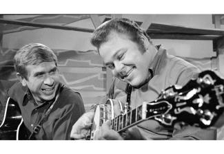 Photo of Roy Clark playing the banjo with Buck Owens holding a guitar and smiling watching Roy play