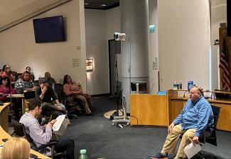 Two individuals sit in chairs acting out a scene with another individual sitting in a chair looking on, in front of an audience of people.