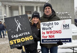 Two SAG-AFTRA New England Local members pose for a photo outside of the New Hampshire State Legislative Building. Both wear SAG-AFTRA hats and winter jackets and hold signs rallying against passing right-to-work legislation in the state.