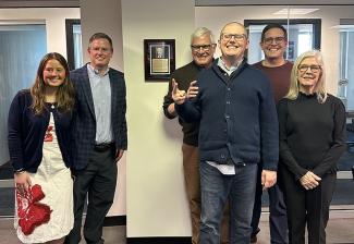 Six close family members of John Henning pose for a photo next to the plaque in the New England Local boardroom officially honoring his legacy, four on the right side and two on the left. 