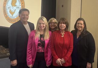 A diverse group of people stand in front of a beige wall. There is the New Jersey State AFL-CIO logo behind them and an American flag to the right.
