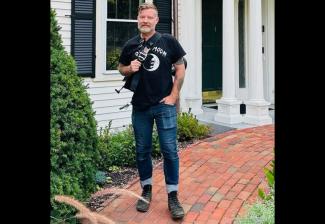 A man wearing blue jeans, boots and a black T-shirt holds a microphone while standing on a red brick sidewalk outside of a white wooden building.