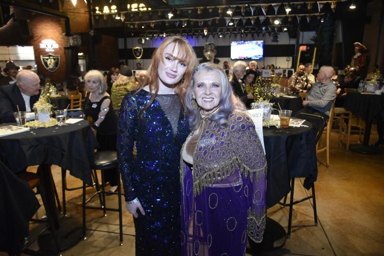 Aniston and Renee stand closely together for a photo. Both wear dresses: Aniston’s dress is a sequined blue and Renee’s is purple with ornate beading. Behind them is a restaurant setting, with seated partygoers.