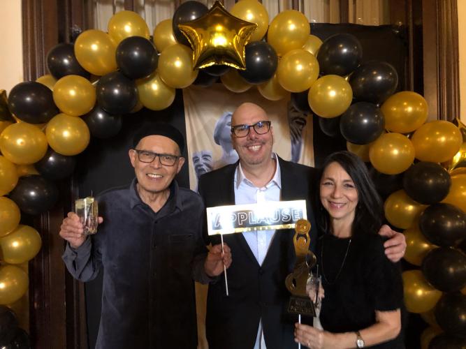 Toliver, Taylor and Gurriere stand close together under a balloon arch. They are dressed in formal party attire. Toliver holds up a drink; Taylor and Gurriere hold up photo booth props, a sign and a paper statuette.