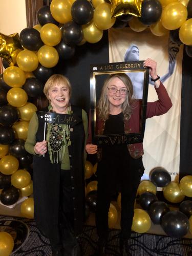 Madden and Howell pose together under a balloon arch. They are dressed in formal party attire: Madden in a black dress with a green scarf; Howell in all-black with a dark red cardigan. They hold up photo booth props. 