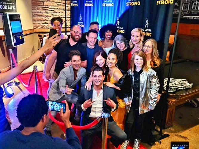 Over a dozen people stand for a group photo in an area that imitates a step-and-repeat. They are dressed in business casual or festive attire, and strike poses for others who take their picture.