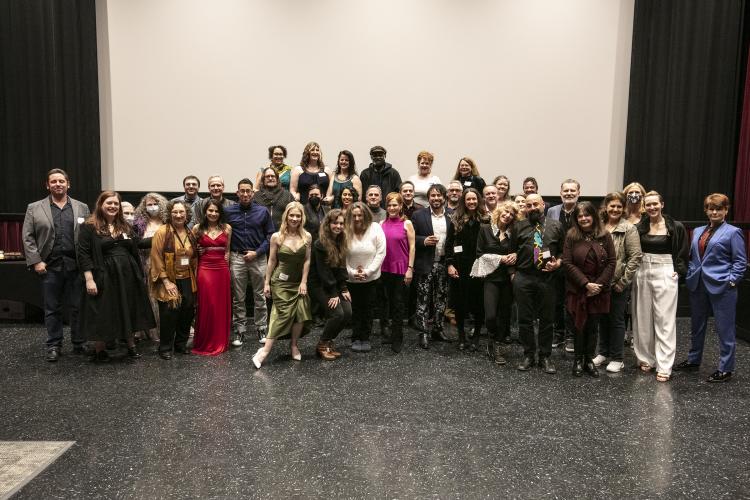 A wide shot of a large group of people arranged in three rows. Those in the third row stand on an elevated platform. All are dressed in festive cocktail or formalwear. Behind them is a blank projection screen bordered by black curtains.
